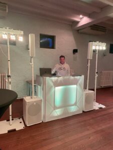 DJ achter verlicht podium, omringd door luidsprekers en verlichting in een interieur met houten vloer.